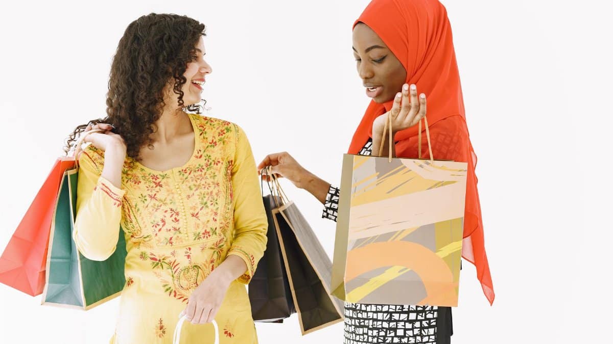 Two women in bright attire smiling and holding shopping bags against a white background.