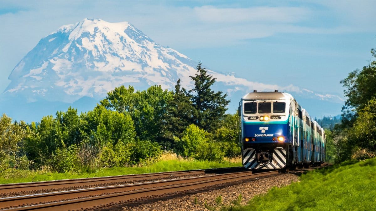 Passenger train traveling with Mount Rainier in the background, lush greenery, bright skies.