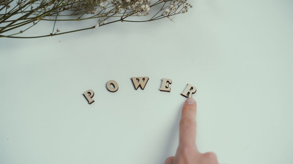 Finger points at wooden letters spelling 'Power' beside delicate flowers on a light background.