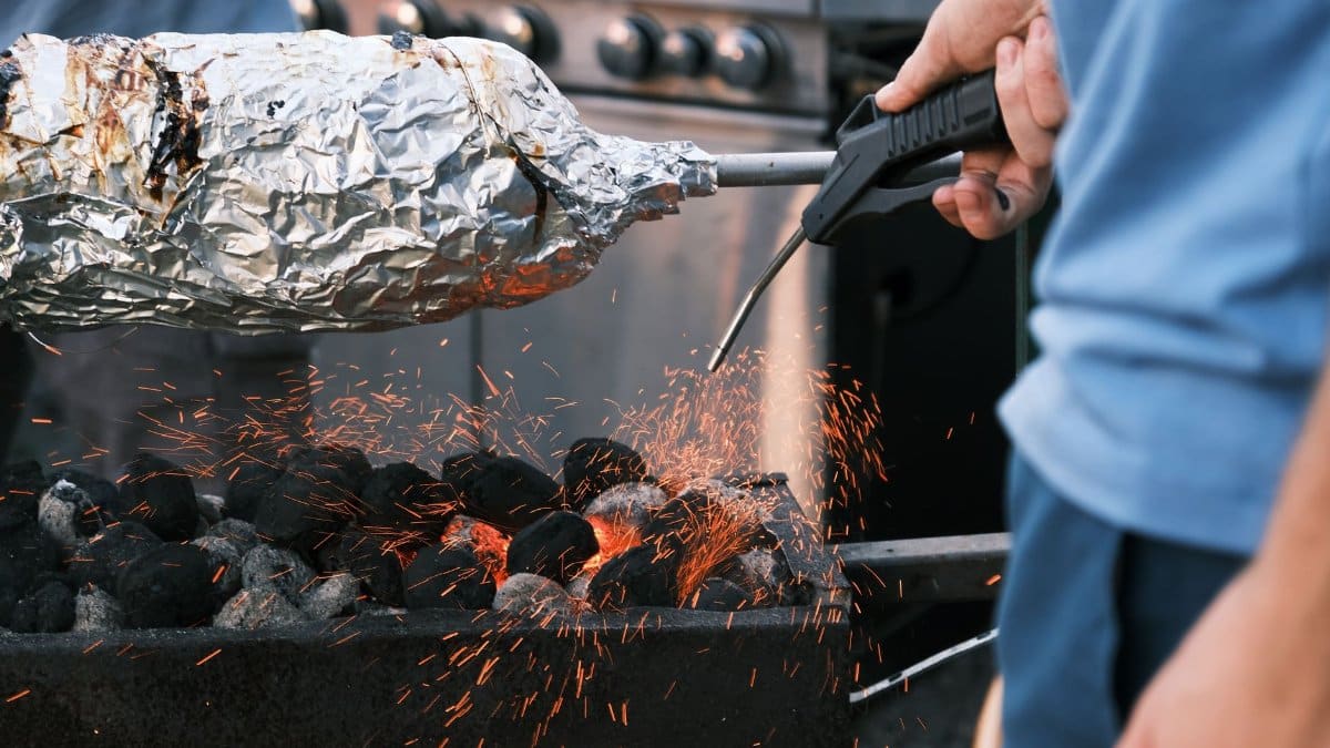 Barbecue cooking with foil-wrapped food over charcoal flames showing flying sparks and intense heat.