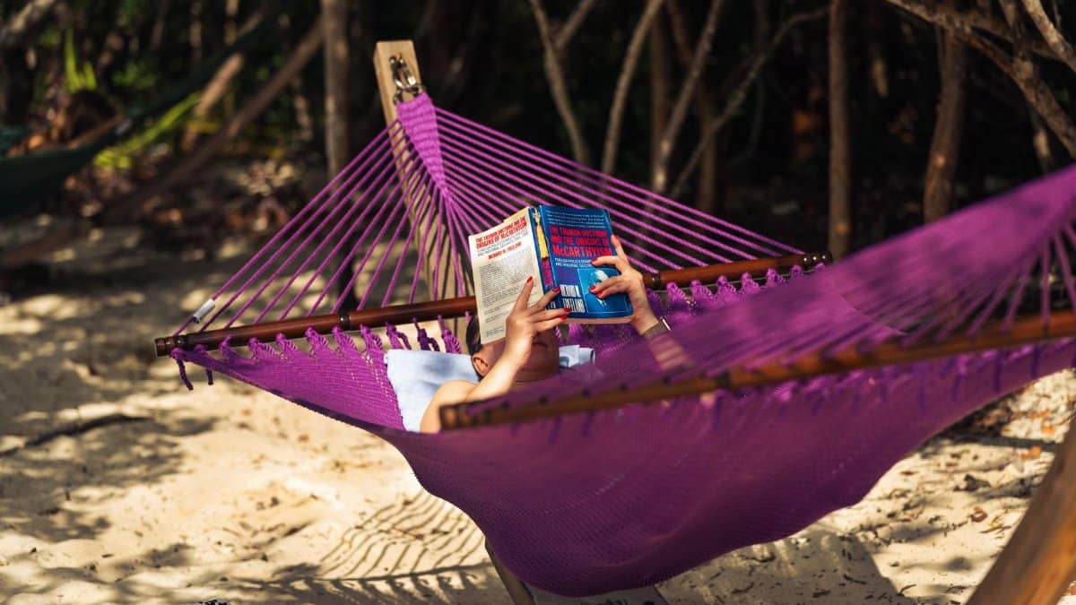 A person enjoys a peaceful read in a hammock under tropical trees on a sandy beach.