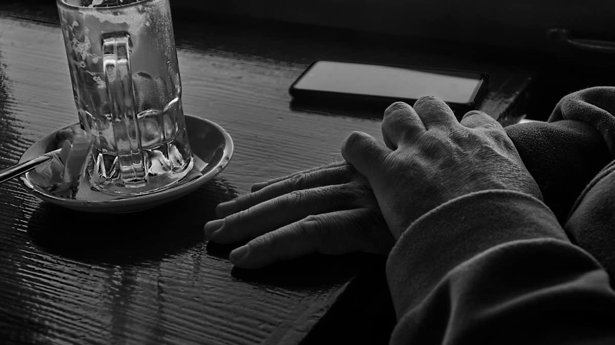 Black and white image featuring hands near a glass mug and cell phone on a table.