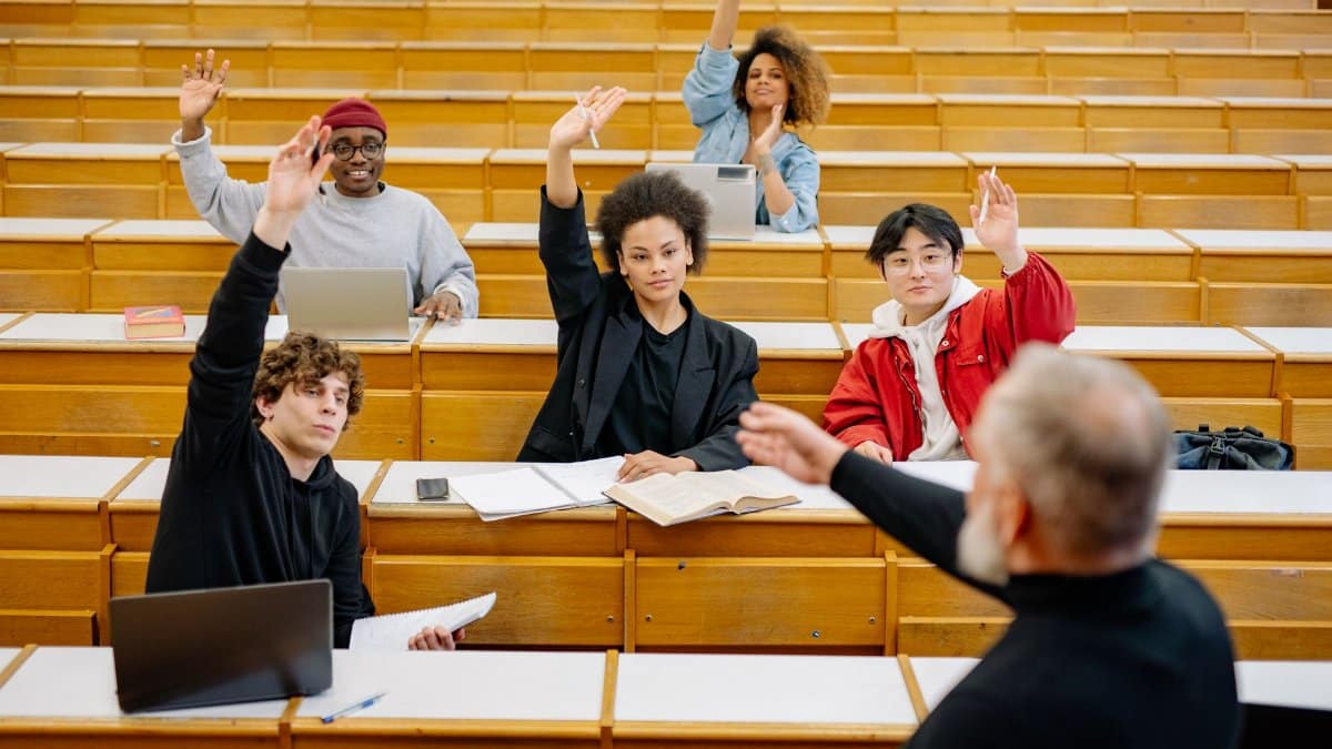 A diverse group of university students participating in a lecture, raising hands to engage with the professor.