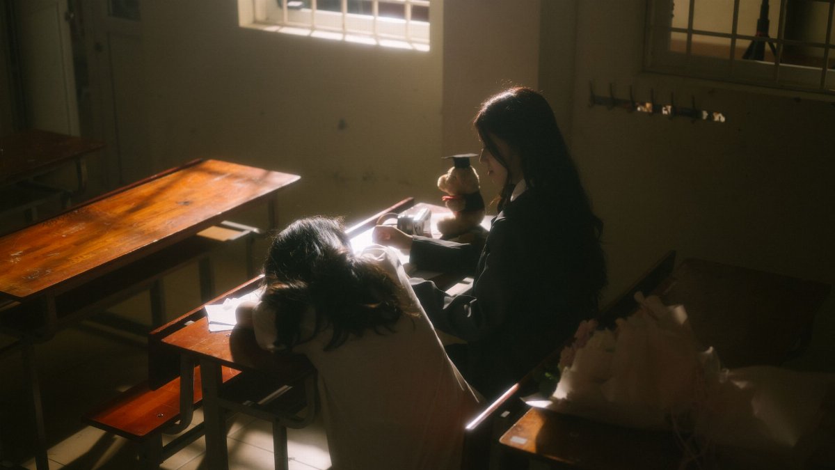 Two students in a dimly lit classroom, one resting on a desk, creating a moody atmosphere.