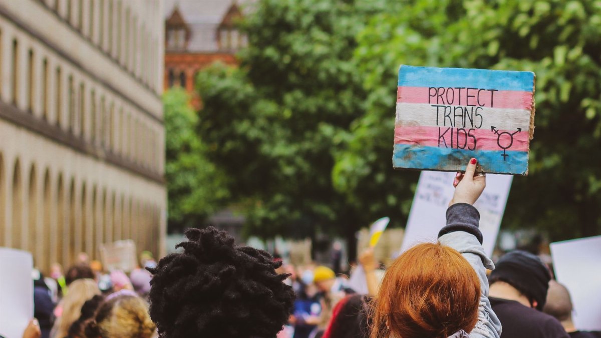 Activists rally outdoors, holding signs to support trans kids' rights. Vibrant urban protest scene.