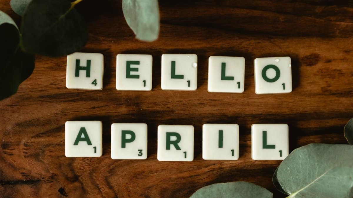 Scrabble tiles arranged to spell Hello April on a wooden surface with green leaves decor.
