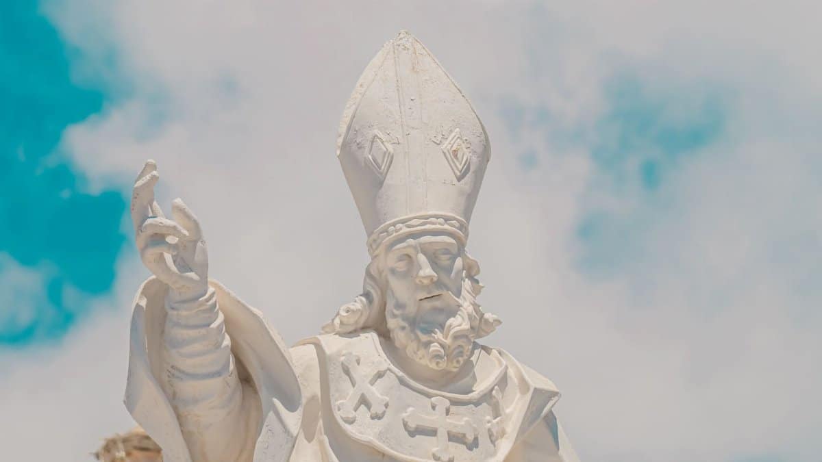 Marble statue of a religious figure dressed as a bishop against a cloud-filled blue sky.