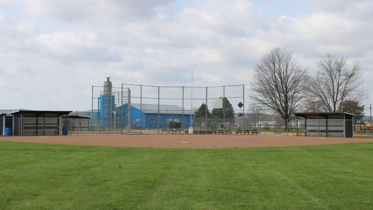 An empty baseball field with dugouts and a blue building in the background under a cloudy sky.