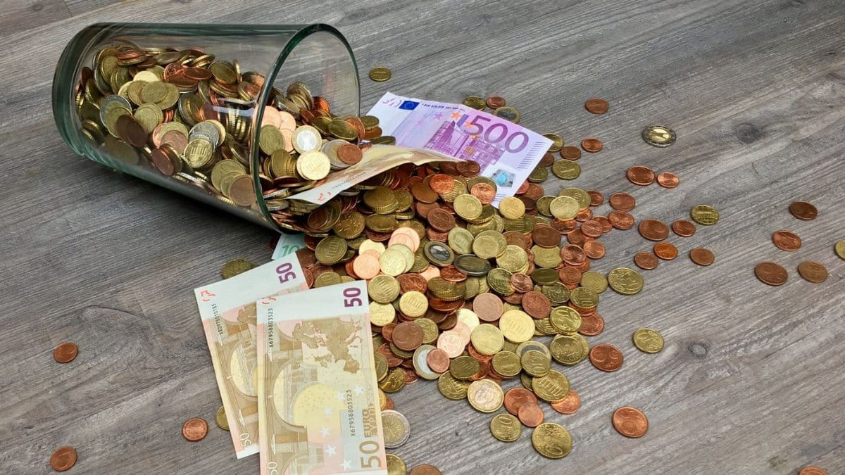 A glass jar with euro coins and banknotes spilled on a wooden surface, representing finance and savings.