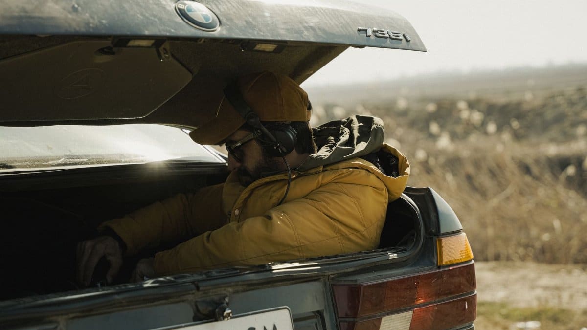 A man wearing headphones sits in the trunk of a car outdoors on a sunny day.