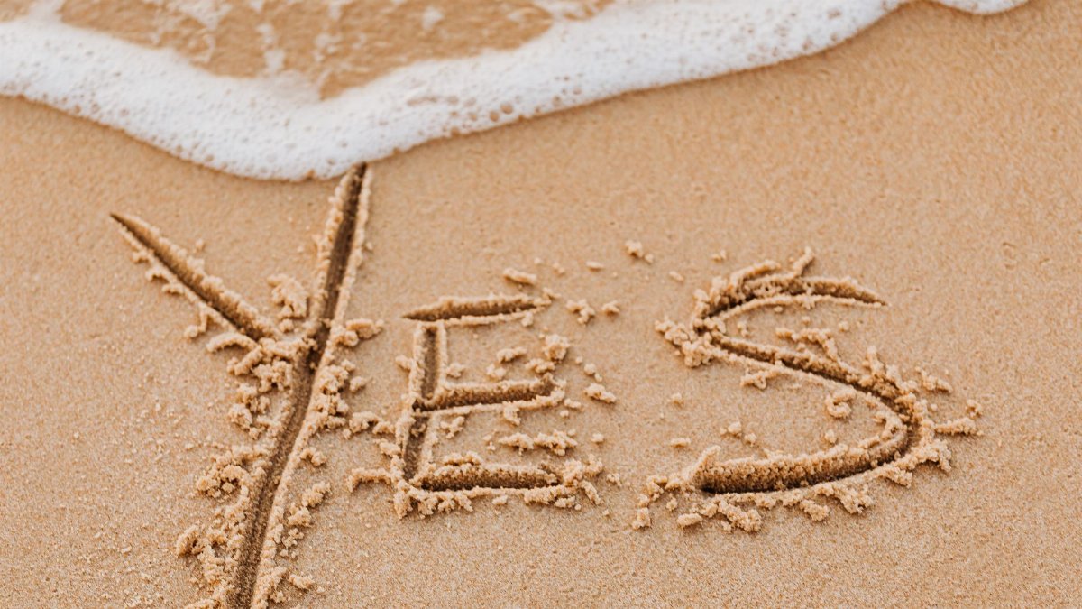 The word 'YES' written in sand with seafoam washing over it.