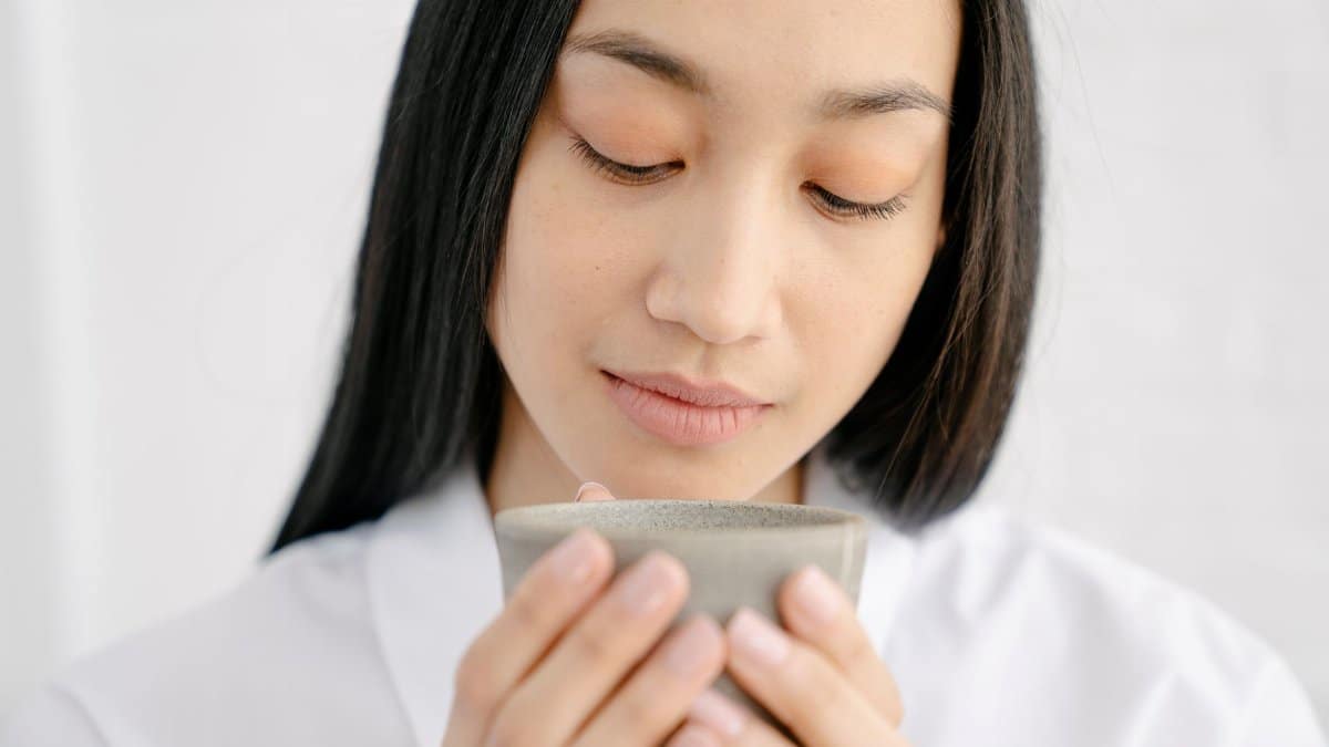 Crop ethnic female with long dark hair smelling aromatic hot beverage in ceramic cup against white background