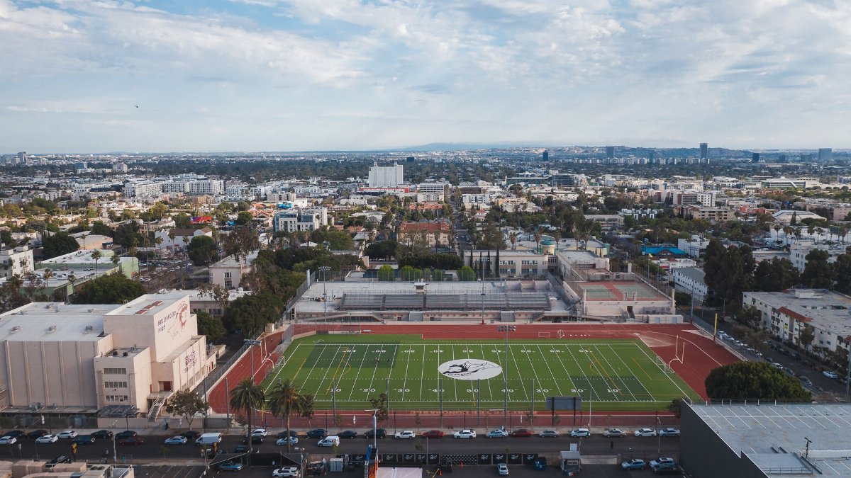 Drone shot capturing an urban football stadium and surrounding cityscape under a bright sky.
