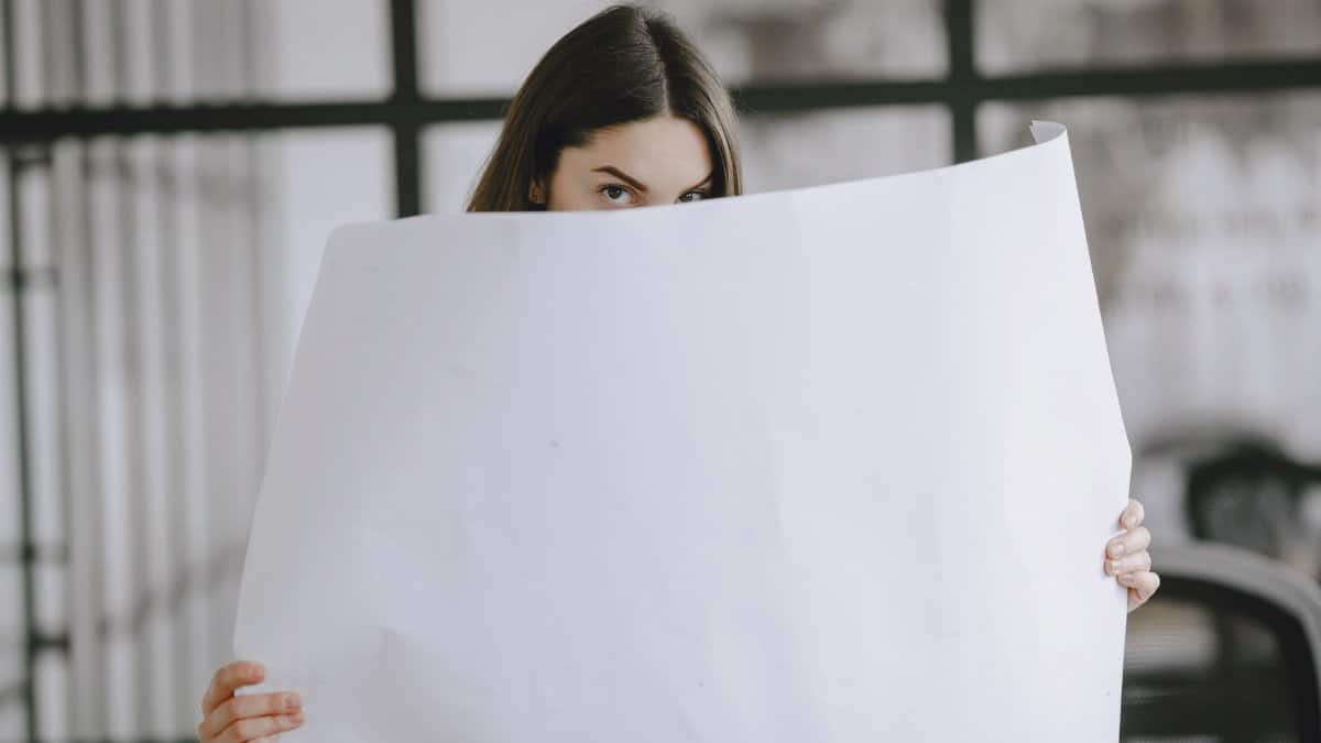 A brunette woman partially hides behind a large white paper sheet indoors, looking directly at the camera.