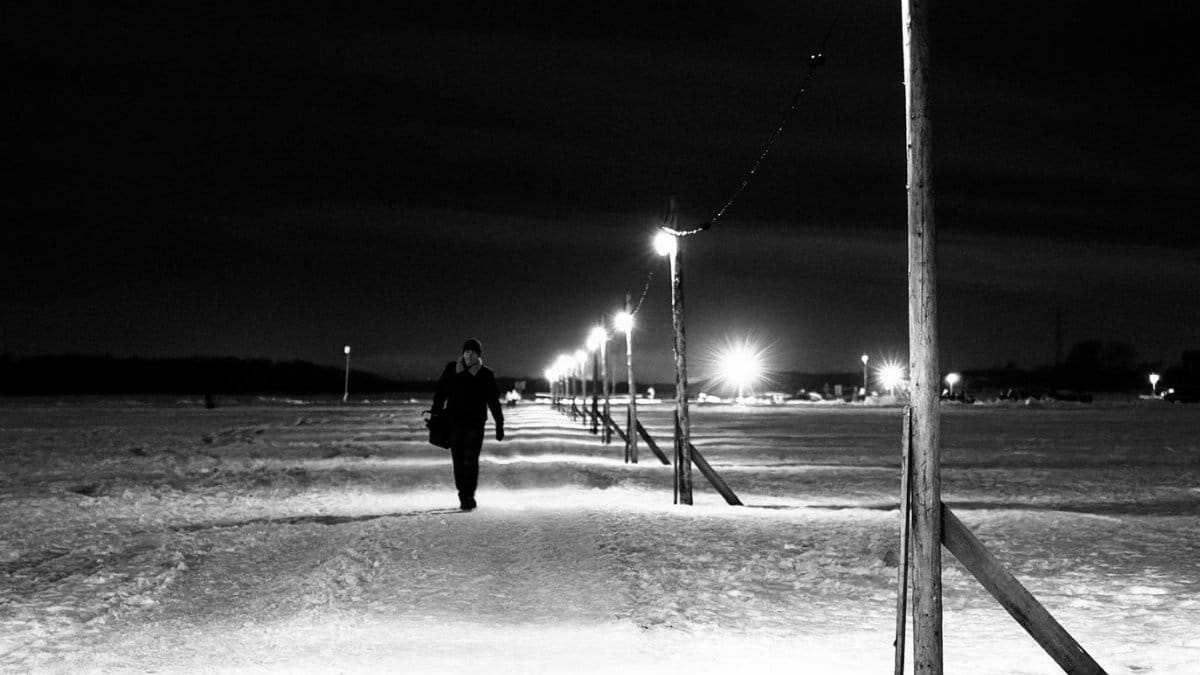 A lone figure walks along a snow-covered path illuminated by street lights at night.