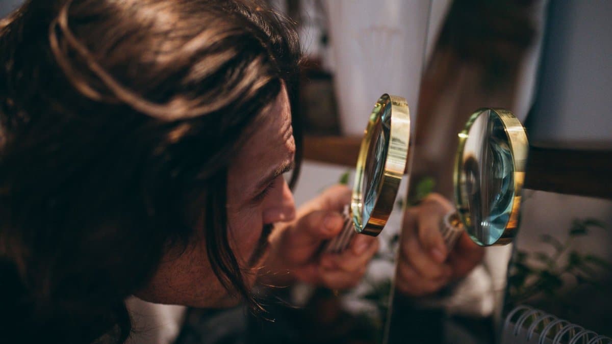 Close-up shot of a man with long hair using a magnifying glass to inspect his reflection in a mirror.