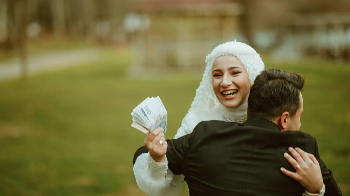 Happy bride in hijab holding money while embracing her groom outdoors.