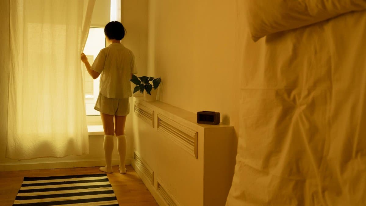 Woman in casual wear opens curtains to let in morning sunlight.