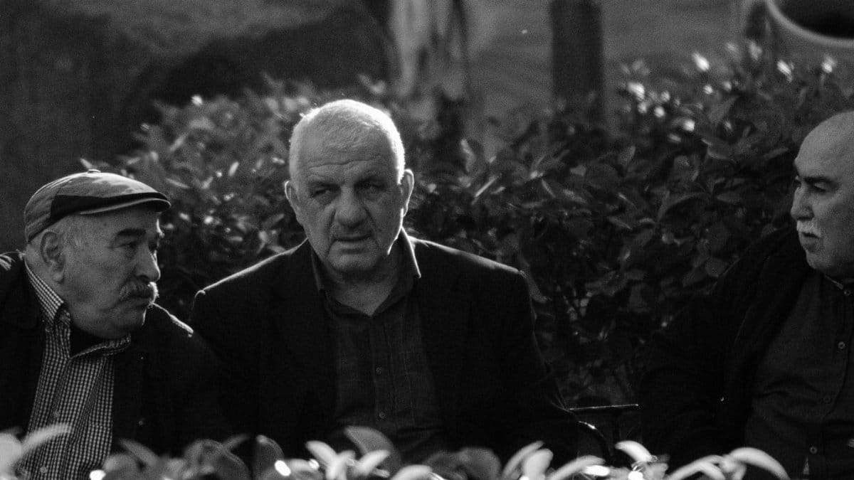 Three elderly men engaged in deep conversation in a natural outdoor setting.