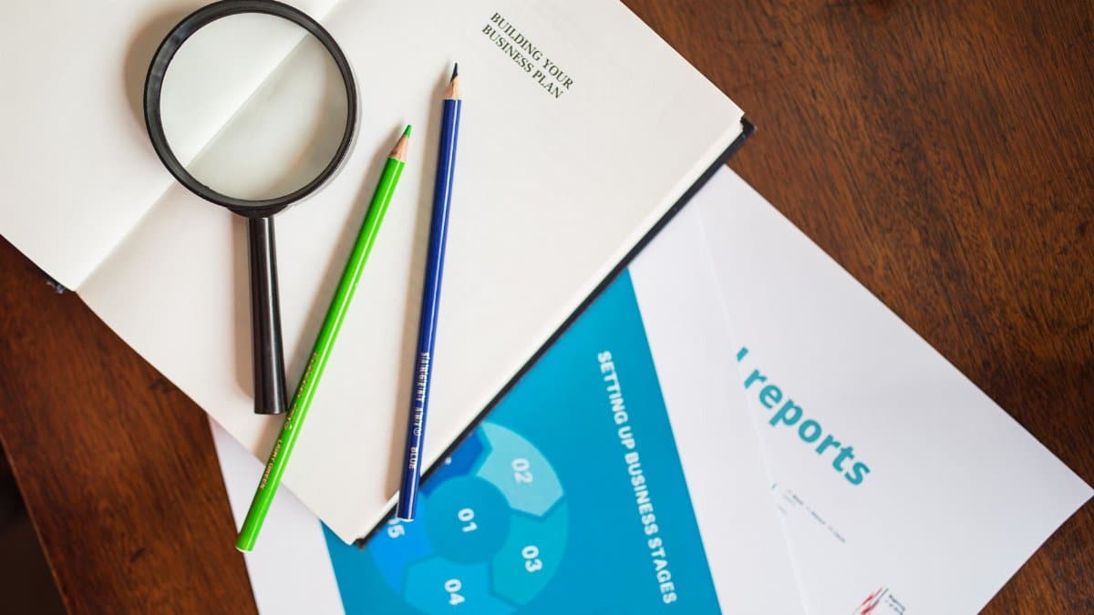 A top view of a magnifying glass, colored pencils, and business reports on a wooden table.