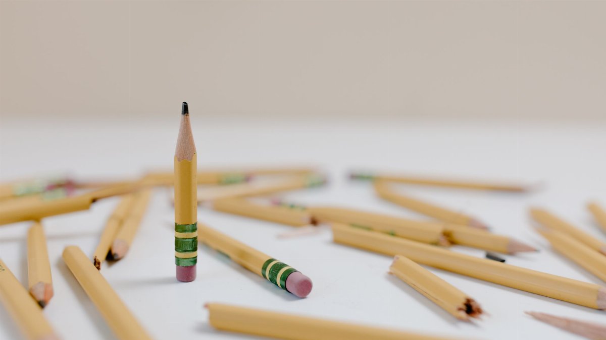 A single upright pencil stands among scattered pencils on a white surface.