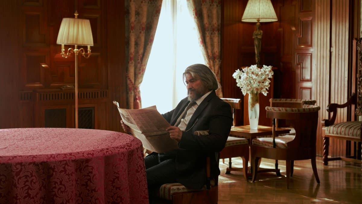 A sophisticated elderly gentleman reads a newspaper in a luxurious, vintage-style room with elegant furnishings.