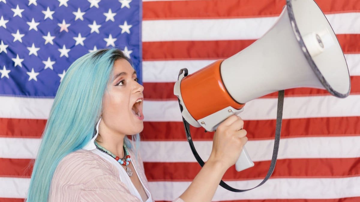 Woman with blue hair shouting through a megaphone in front of an American flag.