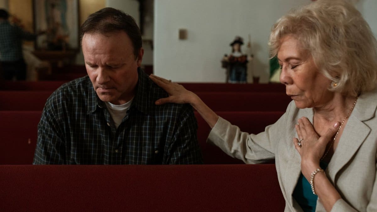 An elderly woman comforts a man praying in a church pew, showing faith and support.