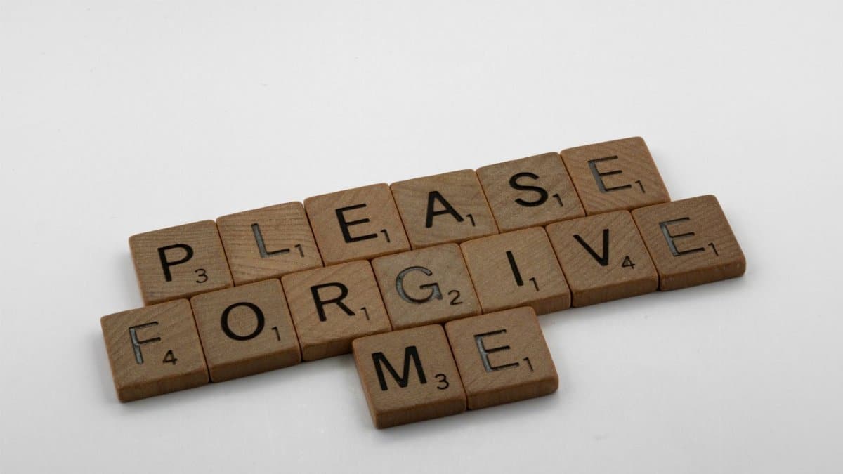 Wooden Scrabble tiles spelling 'Please Forgive Me' on a white surface, emphasizing forgiveness.