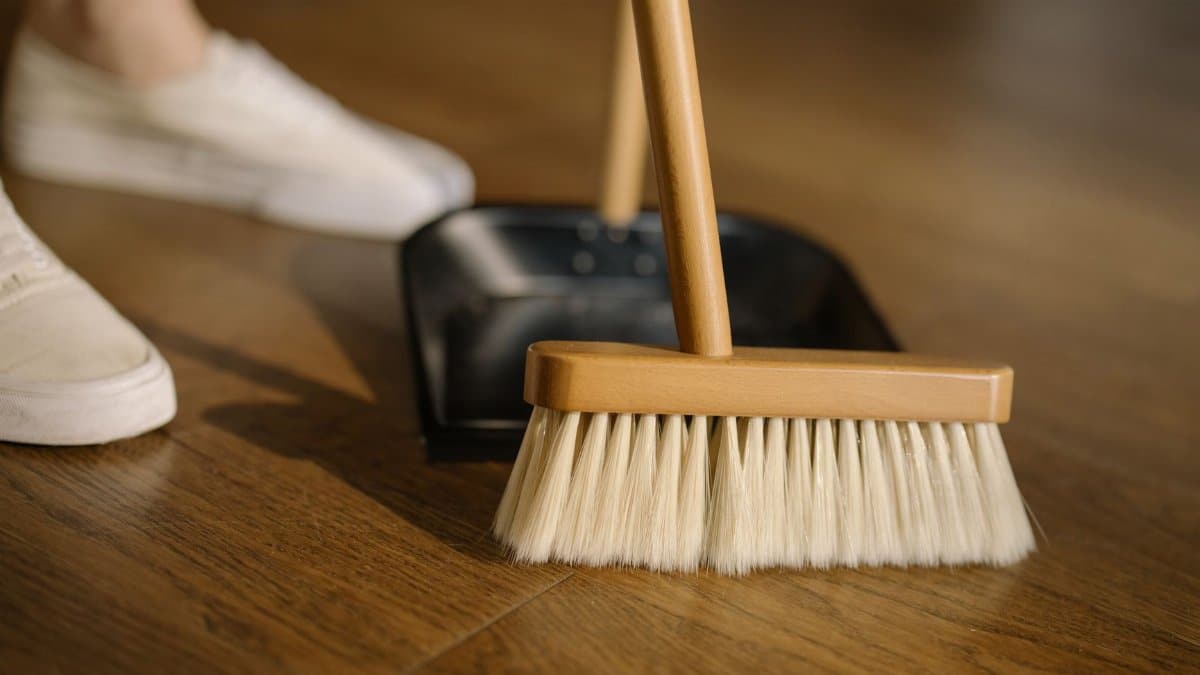 Close-up of a broom and dustpan with white sneakers indoors, representing housekeeping and cleanliness.