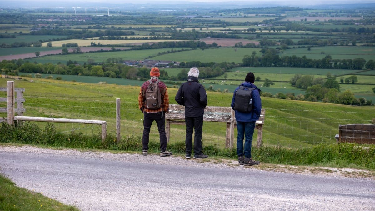 Three hikers with backpacks admire a scenic countryside landscape from an elevated viewpoint.