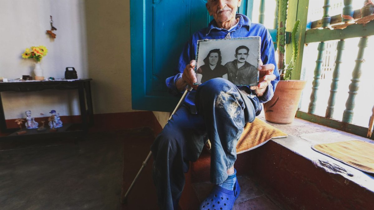 An elderly man sits indoors holding a vintage portrait, reflecting on memories.