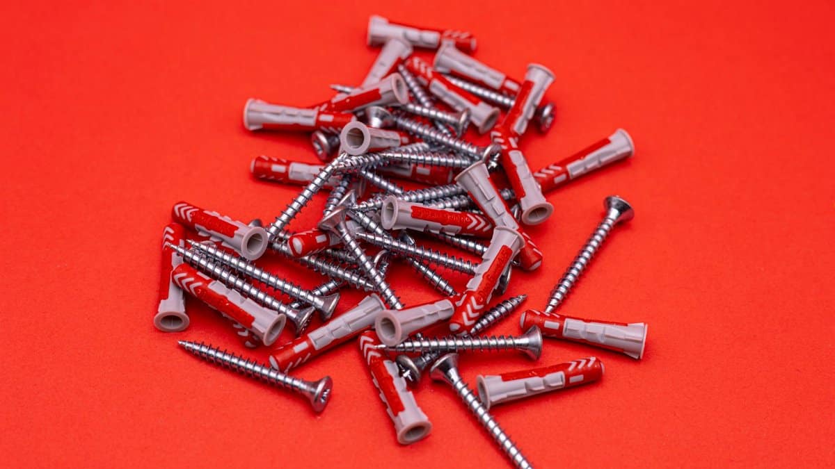 A collection of silver screws and red plastic anchors on a bright red background.