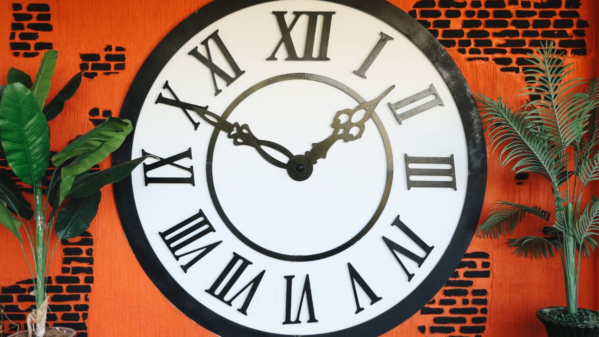 A vintage-style wall clock with Roman numerals against a vibrant orange brick background.