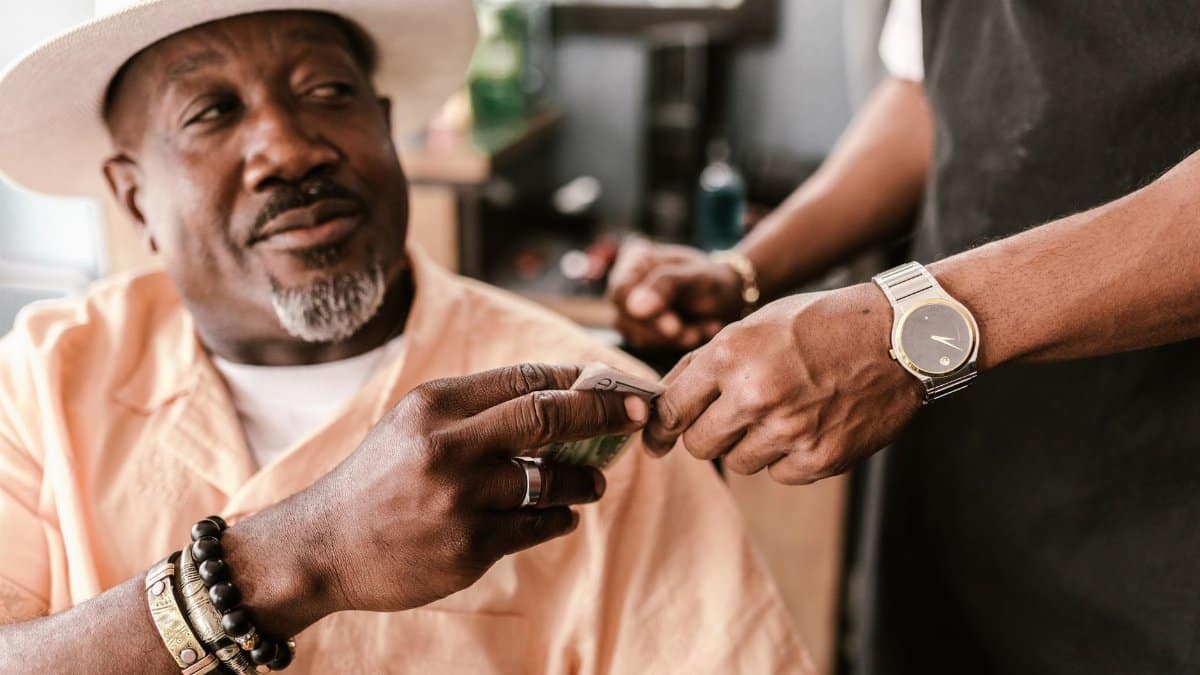 Senior man with a hat giving a tip to a young barber indoors, wearing bracelets and a watch.