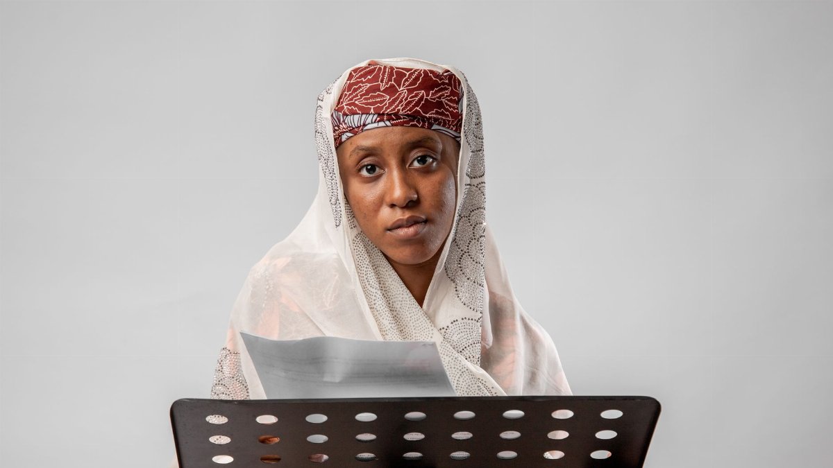A woman in traditional clothing reads from paper on a white background.