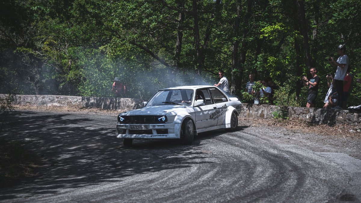 A vintage car drifting around a forest track corner as spectators watch the motorsport event.