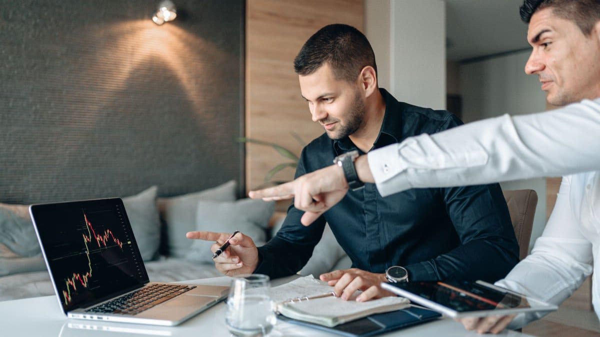 Two businessmen reviewing financial data on a laptop indoors, analyzing market trends.