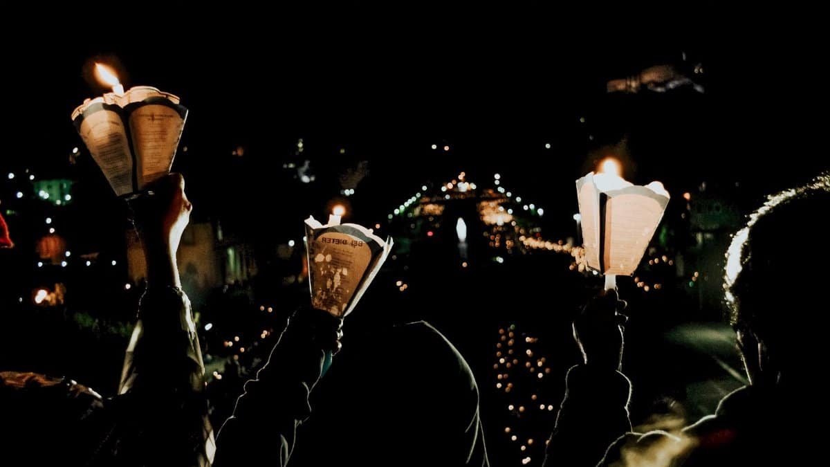 Silhouette of group of people with flaming candles in hands looking at glistening river under black sky at night