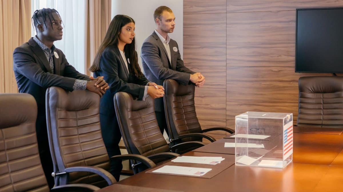 A diverse group of professionals discuss around a ballot box in a conference room.