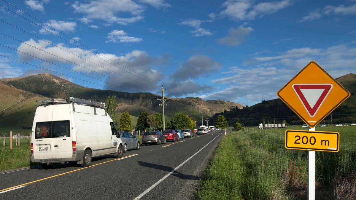 Traffic lined up on a rural road with a yield sign against a mountain backdrop.