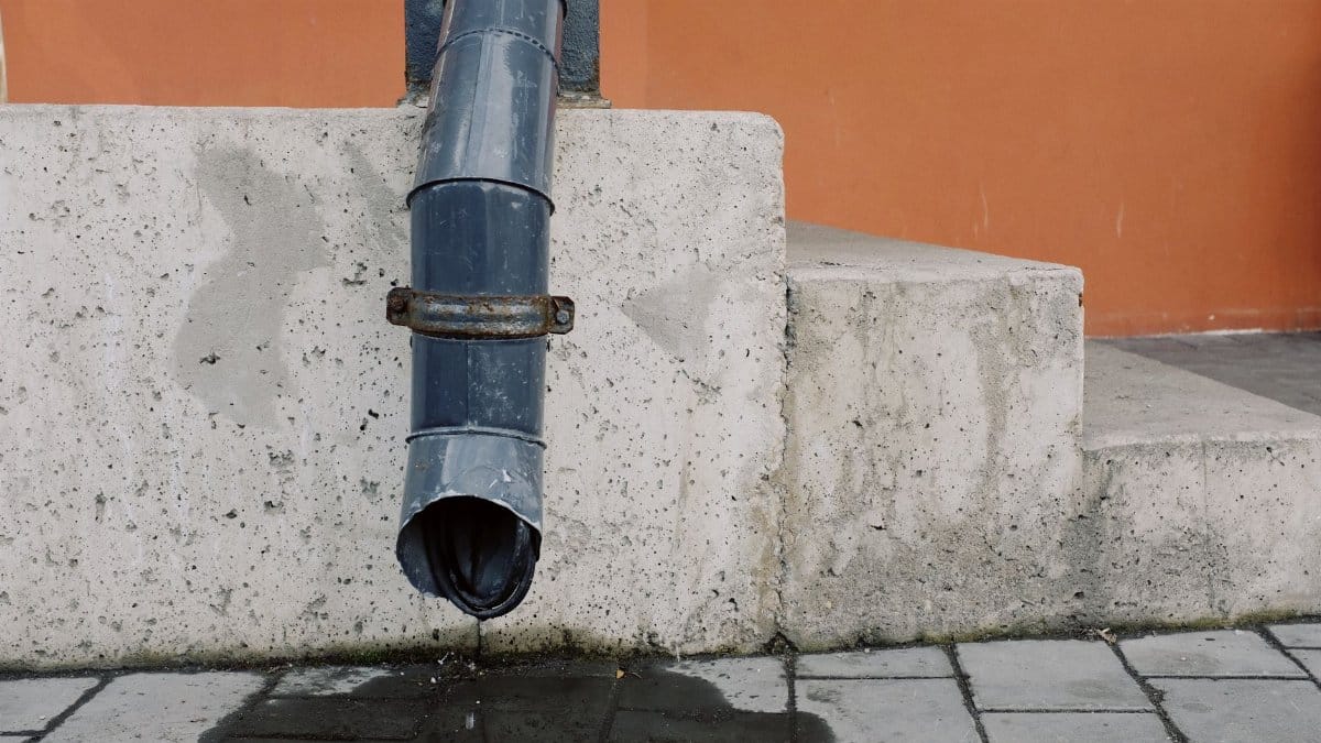 Metal drain pipe with water drops located on red wall near concrete stairs of modern building on street in city