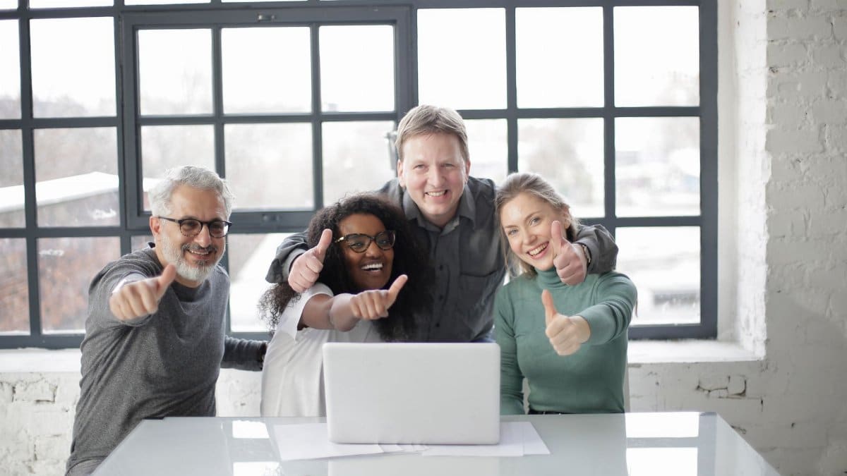 Cheerful diverse coworkers of different ages working on laptop on project while showing thumb up gesture and smiling when looking at camera