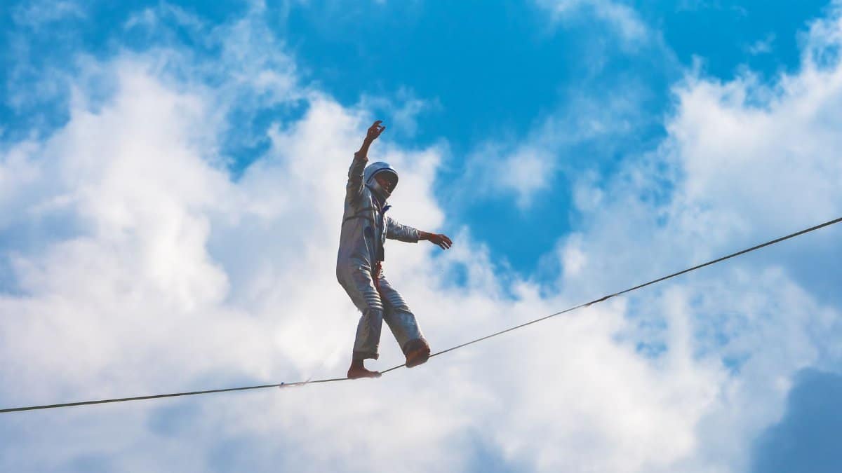 A performer balances on a high wire against a bright blue sky and clouds.