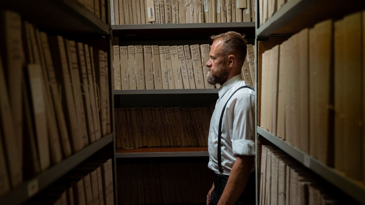 A man in formal attire and suspenders stands in an archive surrounded by documents.