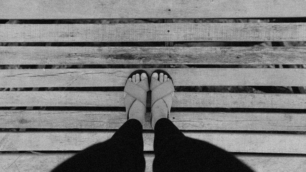 A first person perspective of feet standing on a wooden bridge in black and white.