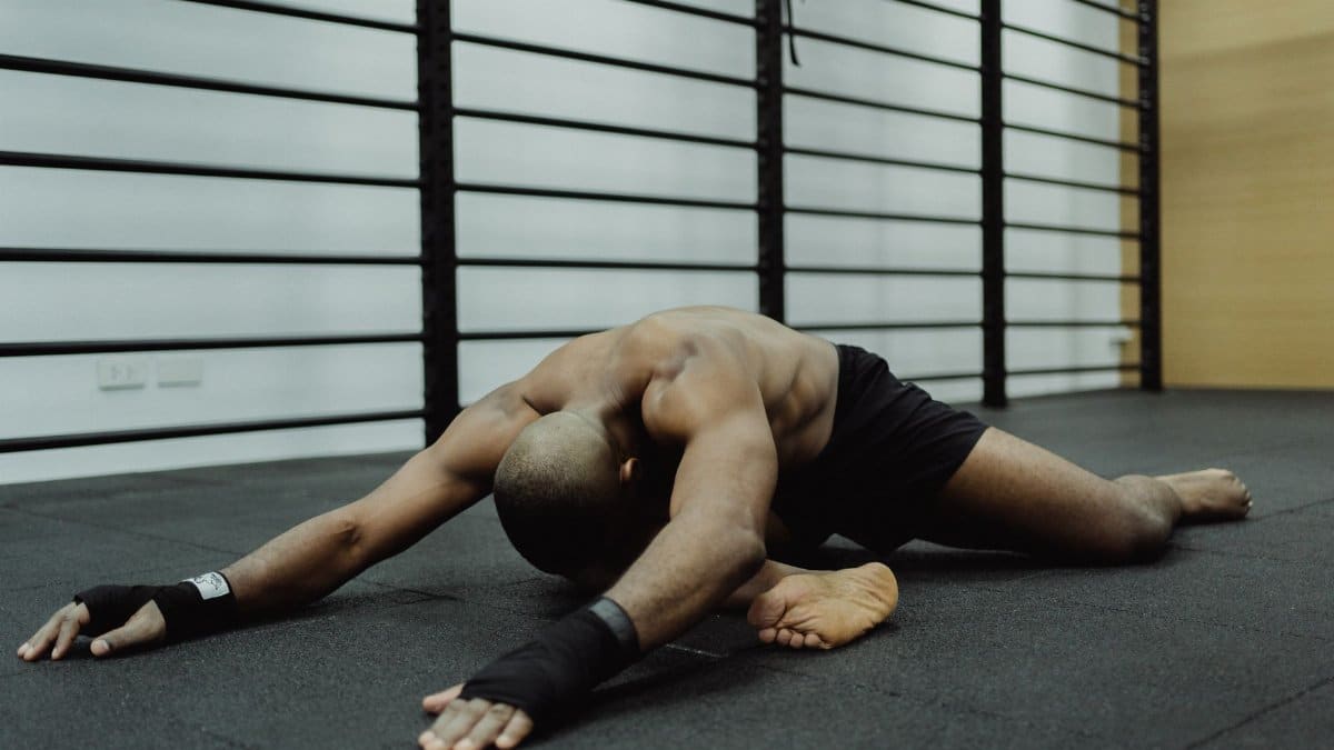 A shirtless man stretches on a black mat in an indoor gym with metal bars.