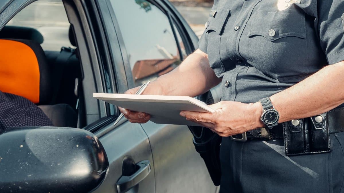 A police officer in uniform writes a ticket to a driver through the car window on a sunny day.