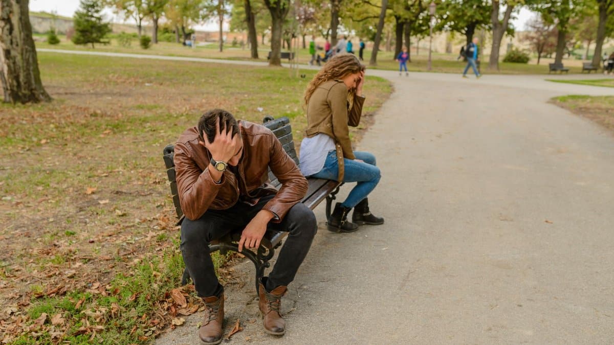 Distraught couple sitting apart on a park bench, facing away from each other, in a public park setting.