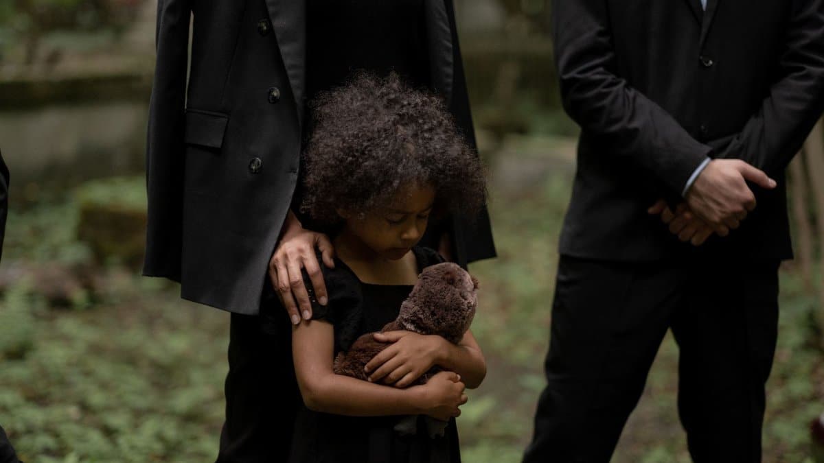 A child holds a plush toy during a somber outdoor funeral service.